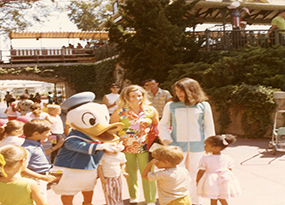 Disney character Donald Duck greats a few of its young fans.  Two mothers stand by and watch their children while other bystanders look on.